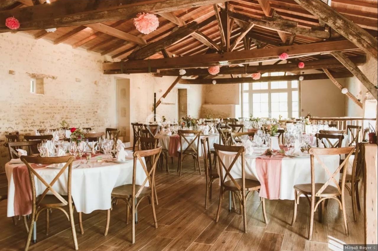 Salle de réception lumineuse avec tables rondes décorées de nappes blanches et chaises en bois, sous une charpente apparente.
