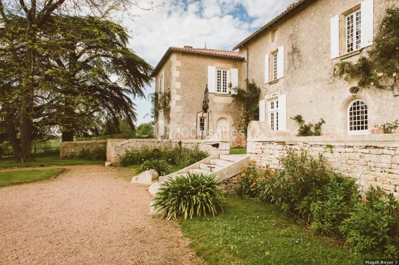 Façade d'une maison en pierre avec escalier, entourée de verdure et d'un jardin fleuri sous un ciel partiellement nuageux.
