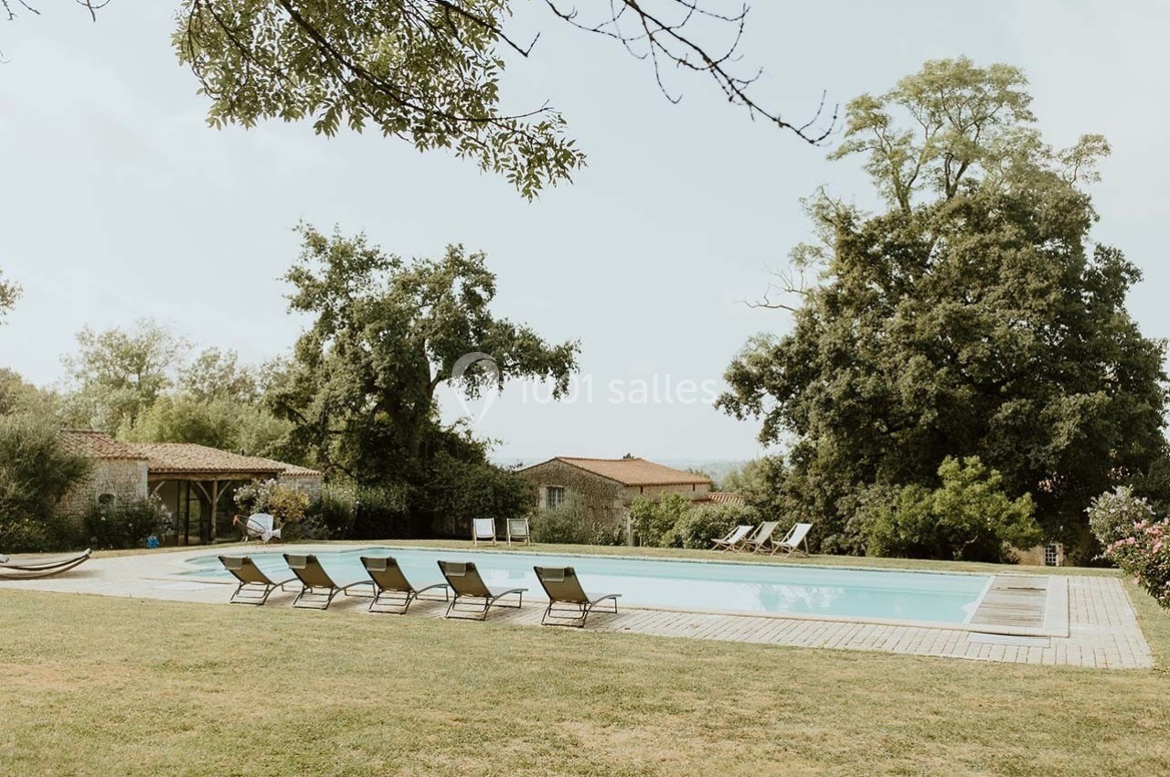 Piscine extérieure entourée de chaises longues, pelouse et arbres, avec des bâtiments en pierre à l'arrière-plan.