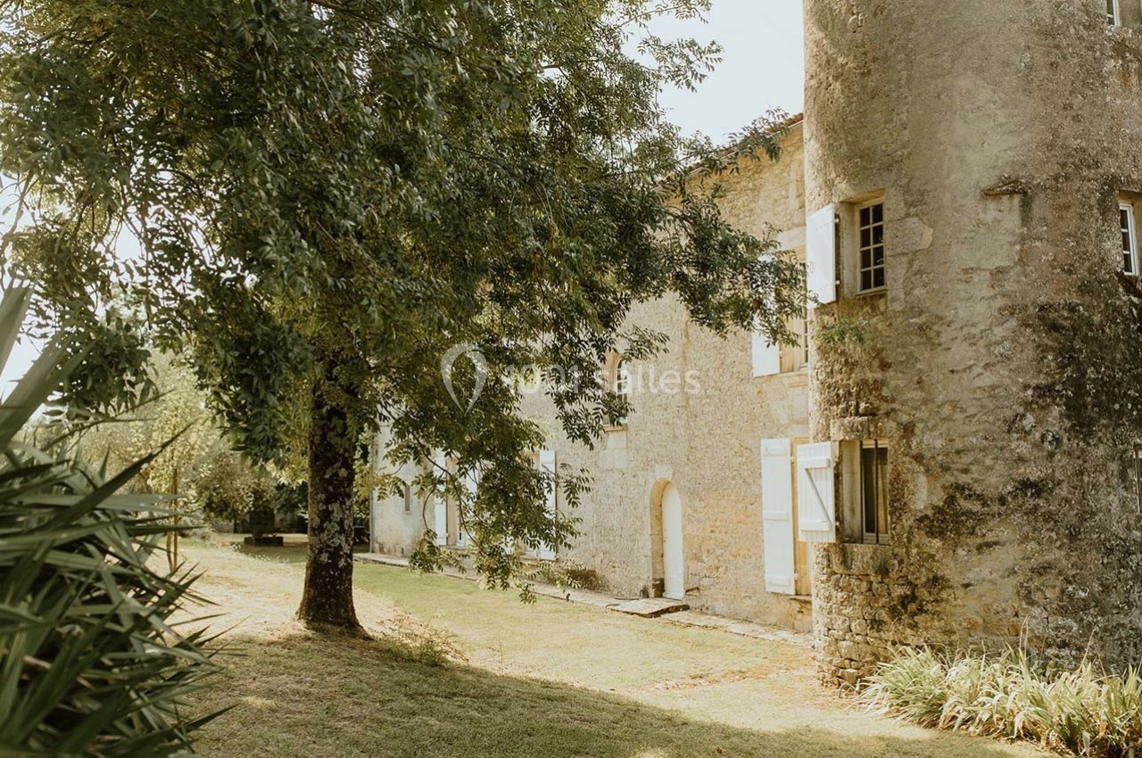 Façade en pierre d'une maison ancienne avec volets blancs, entourée d'arbres et d'un jardin verdoyant.