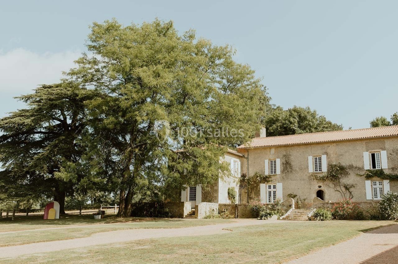 Grande maison en pierre avec volets blancs, entourée d'arbres et d'un chemin de gravier sous un ciel dégagé.