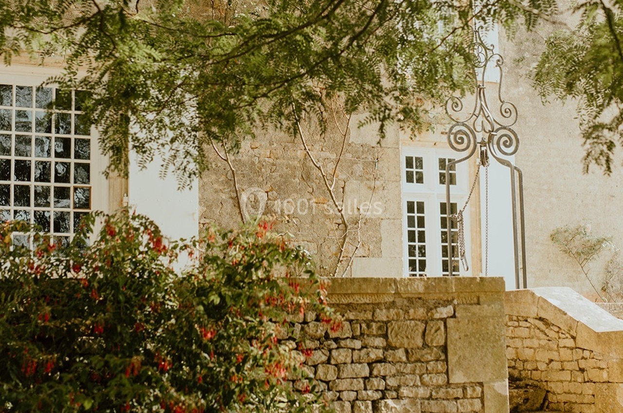 Façade en pierre avec fenêtres blanches, entourée de verdure et d'un mur en pierre, sous des arbres.