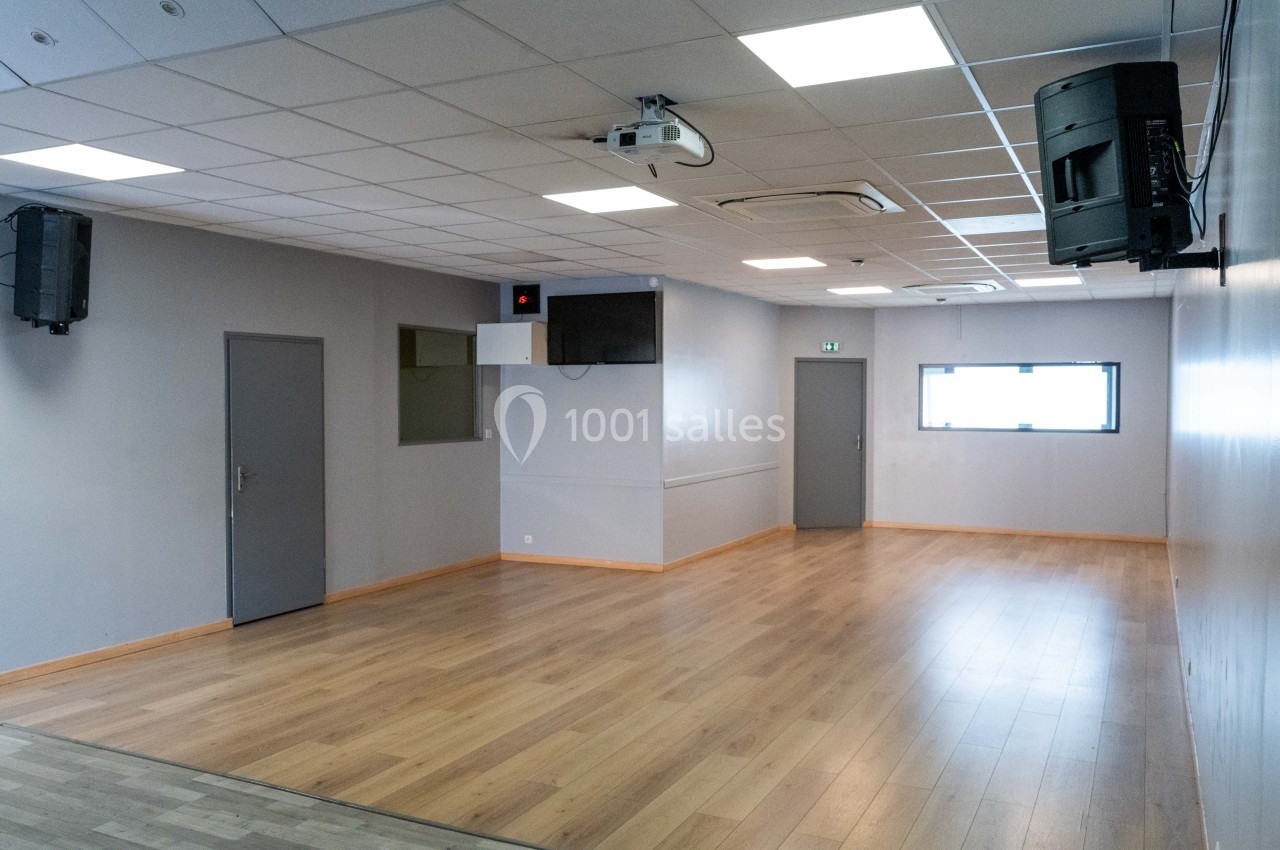 Salle vide avec parquet, murs clairs, plafond à dalles, éclairage intégré, fenêtres et équipements audiovisuels fixés.