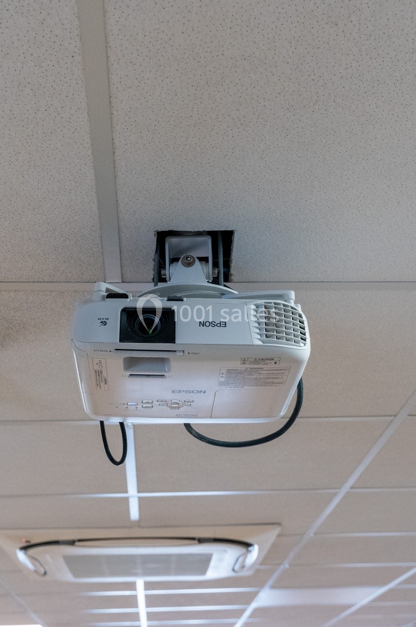 Projecteur fixé au plafond d'une salle, orienté vers le bas, avec un câble d'alimentation visible.