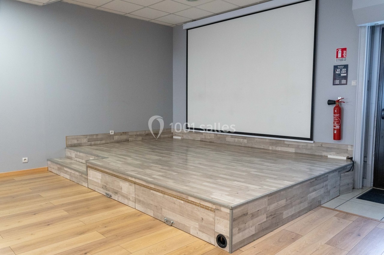 Petite scène en bois clair avec écran de projection blanc en arrière-plan, dans une salle au sol en parquet.
