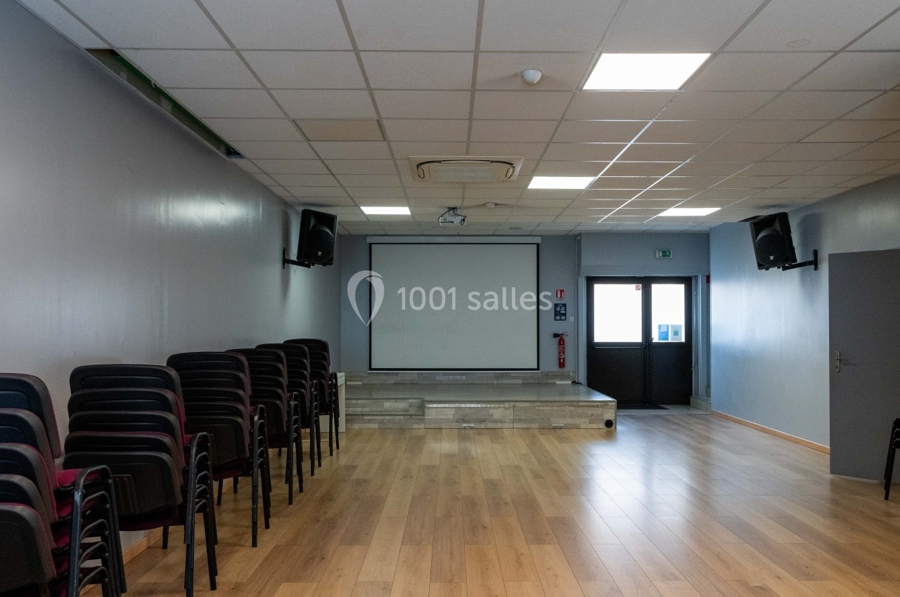 Salle vide avec parquet, écran de projection, chaises empilées sur le côté et éclairage au plafond.