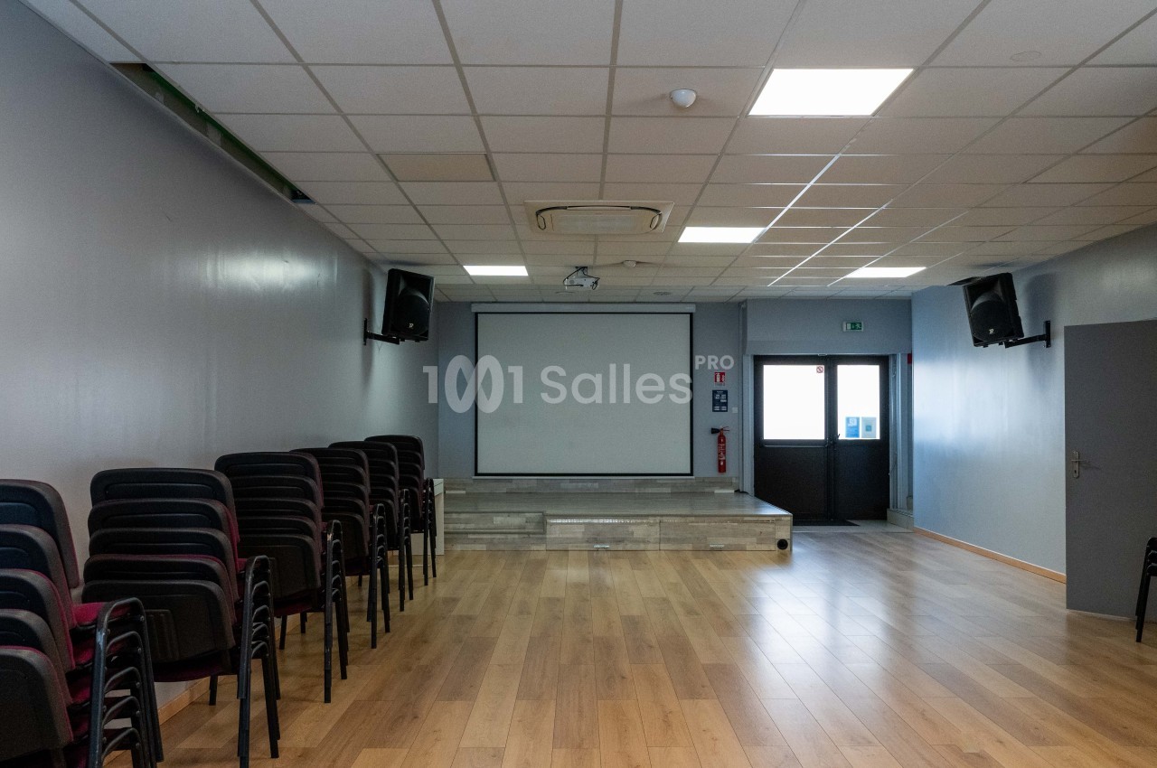 Salle vide avec parquet, écran de projection, chaises empilées sur le côté et éclairage au plafond.