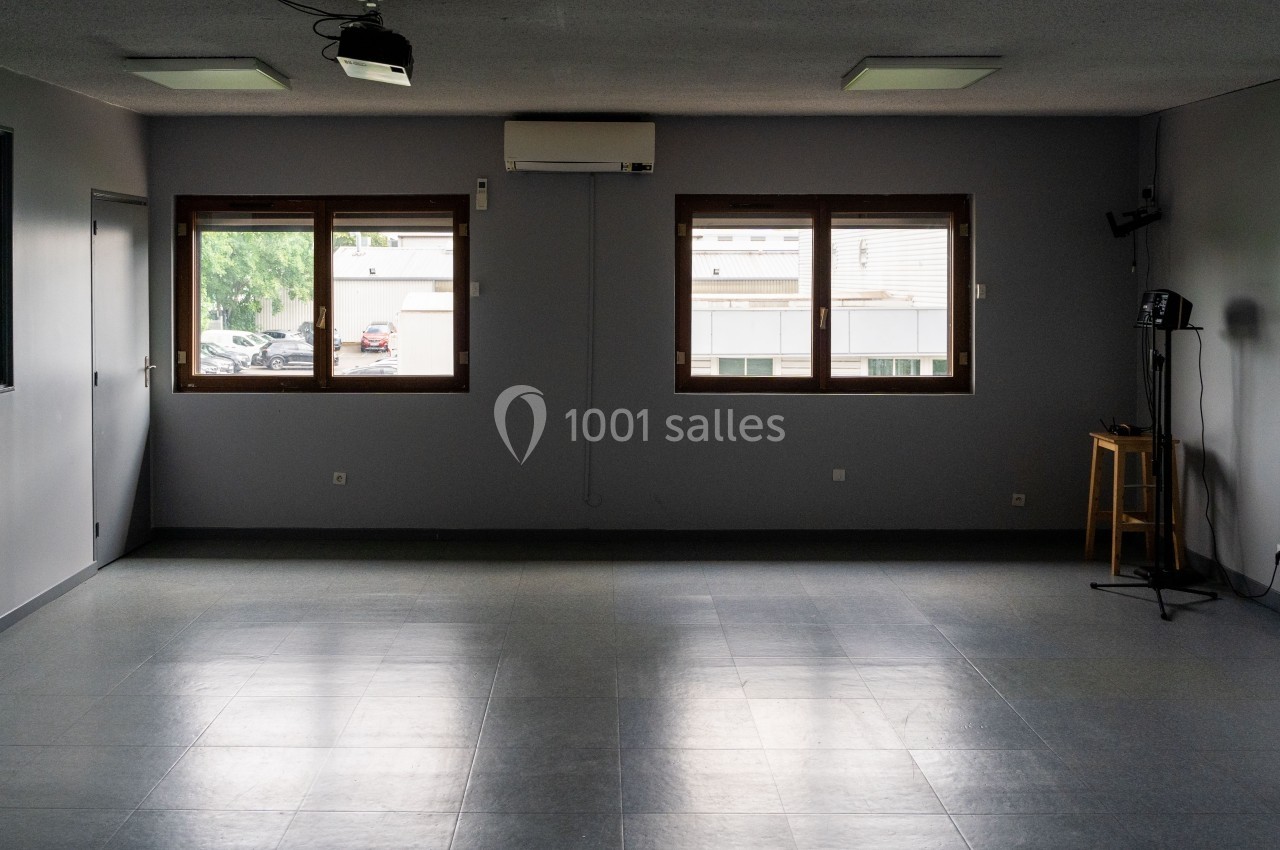Salle vide avec sol carrelé, murs gris, trois fenêtres laissant entrer la lumière naturelle et quelques équipements.