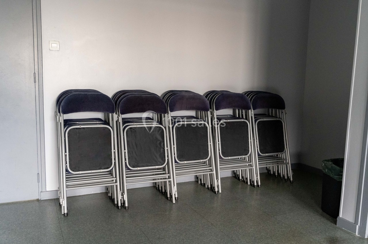 Chaises pliantes empilées contre un mur gris dans une pièce avec un sol en carrelage et une poubelle noire à droite.