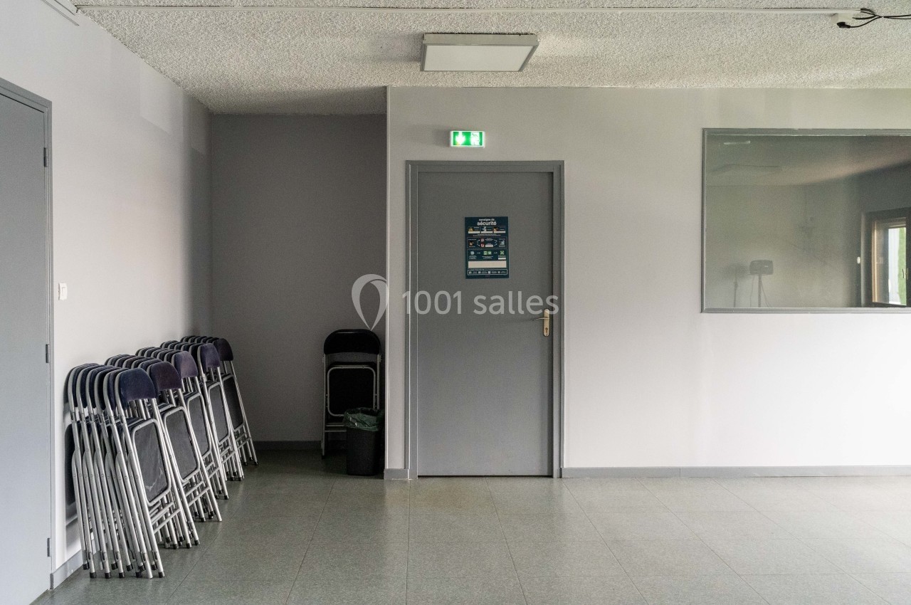 Un couloir avec des chaises empilées à gauche, une porte grise au centre et une fenêtre à droite.