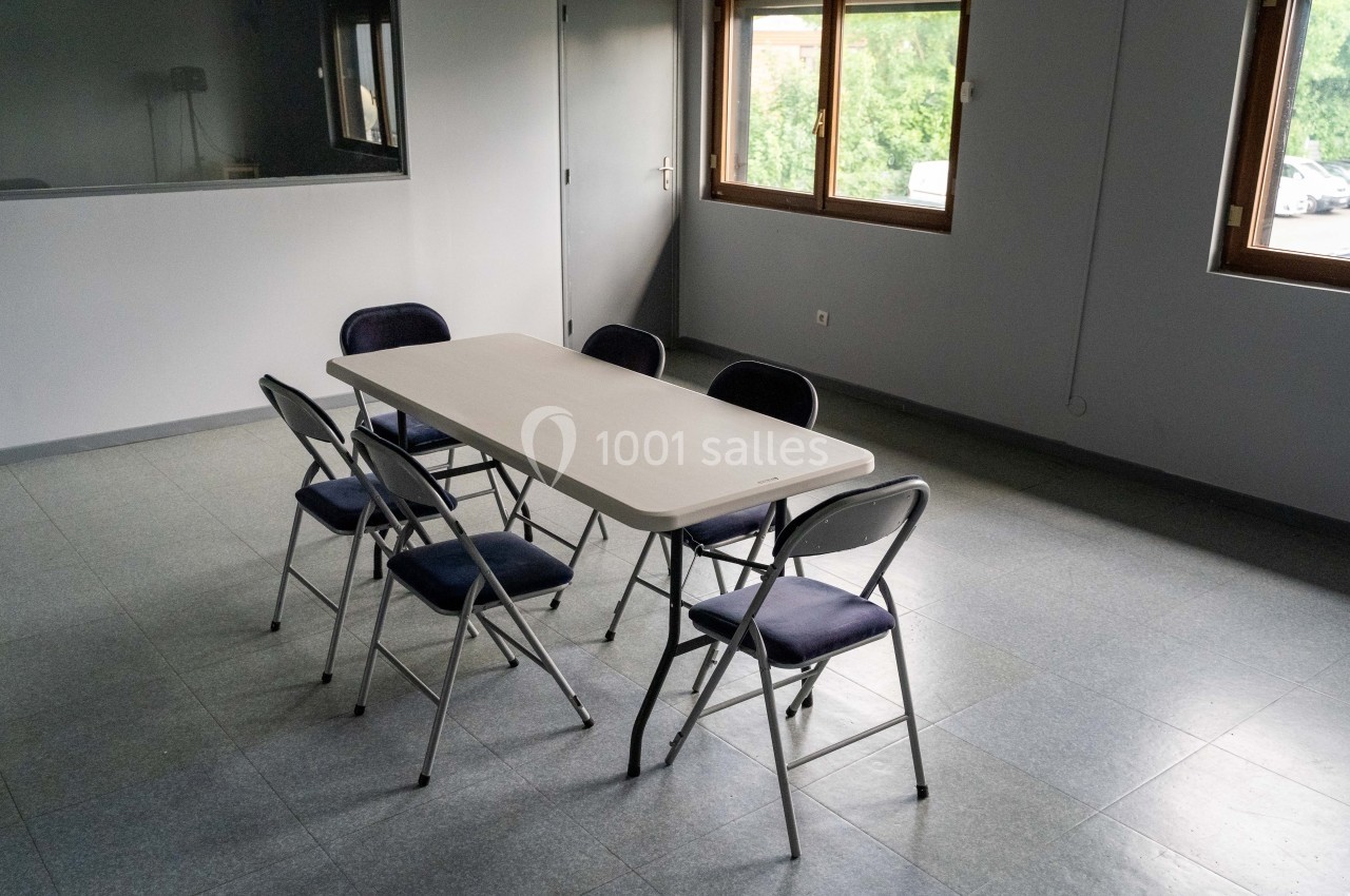 Salle vide avec une table rectangulaire blanche entourée de six chaises pliantes, éclairée par des fenêtres.