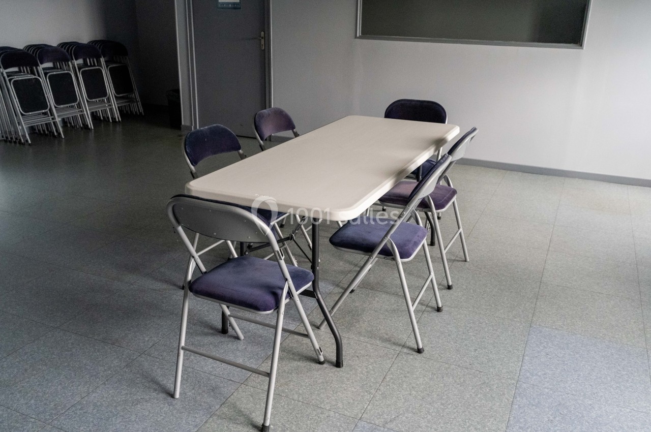 Table rectangulaire blanche entourée de six chaises pliantes dans une pièce aux murs clairs, sol en carrelage.