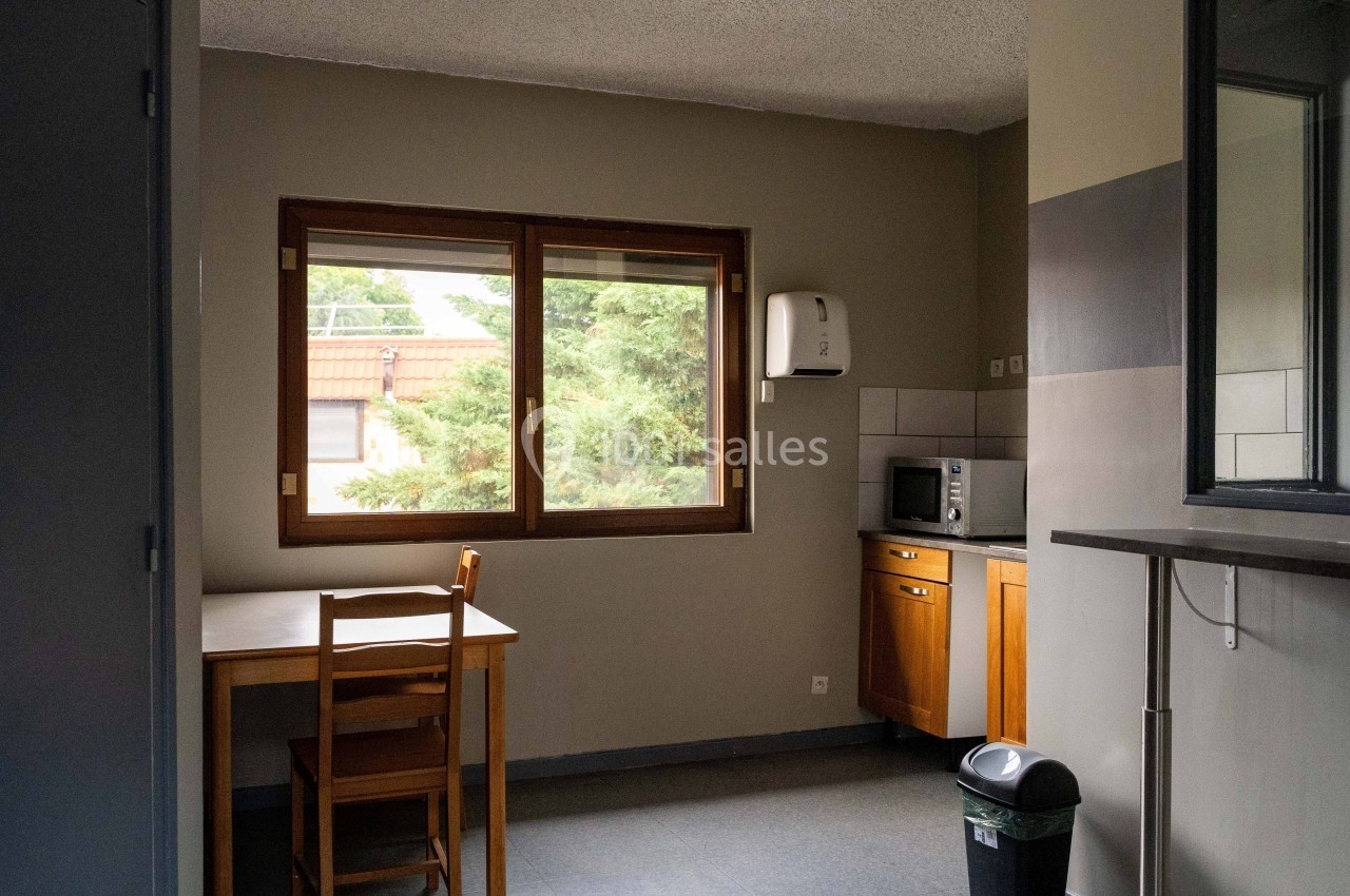 Cuisine simple avec table, chaise, micro-ondes, poubelle et fenêtre donnant sur un extérieur arboré.