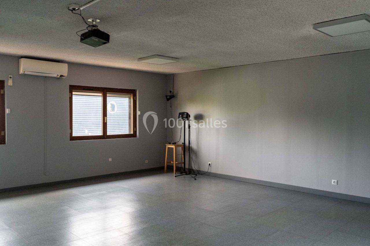 Salle vide avec murs gris, sol carrelé, fenêtres à gauche, projecteur au plafond et trépied avec équipement à droite.