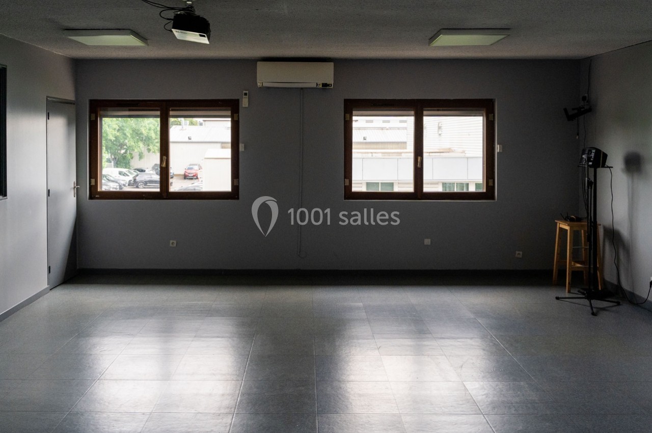 Salle vide avec sol carrelé gris, murs sombres, trois fenêtres laissant entrer la lumière naturelle et quelques équipements.