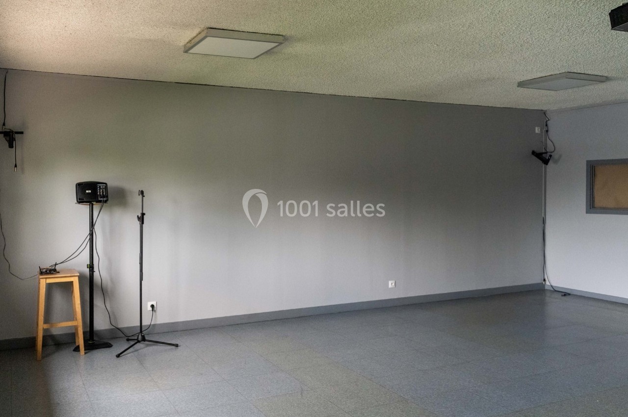 Salle vide avec sol carrelé, murs gris, plafond avec éclairage, un tabouret, un micro sur pied et une enceinte.