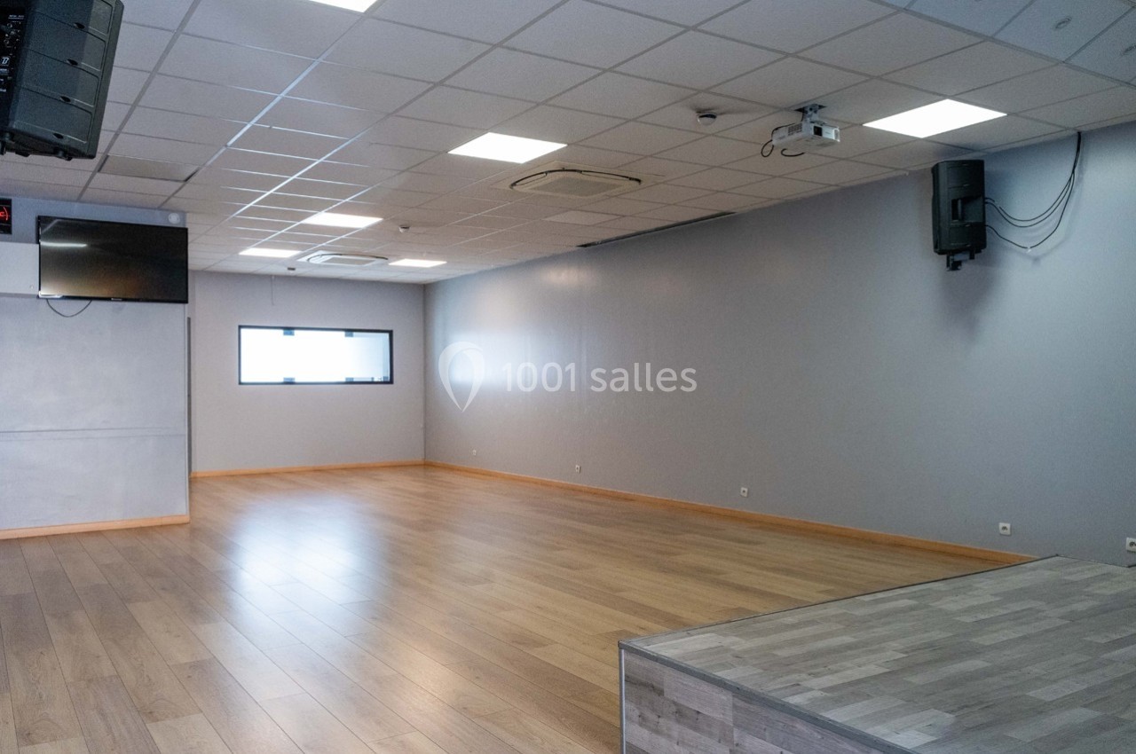 Salle vide avec parquet, murs gris, plafond à dalles blanches, éclairage intégré et estrade en bois au premier plan.