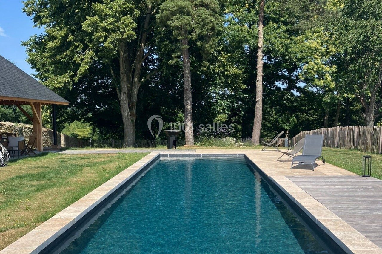 Piscine rectangulaire entourée de pelouse, terrasse en bois et chaises longues, bordée d'arbres en arrière-plan.