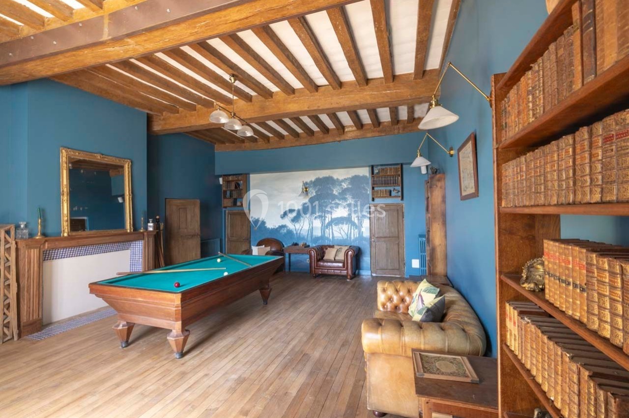 Salle avec un billard, murs bleus, poutres apparentes, bibliothèque en bois et canapé en cuir.