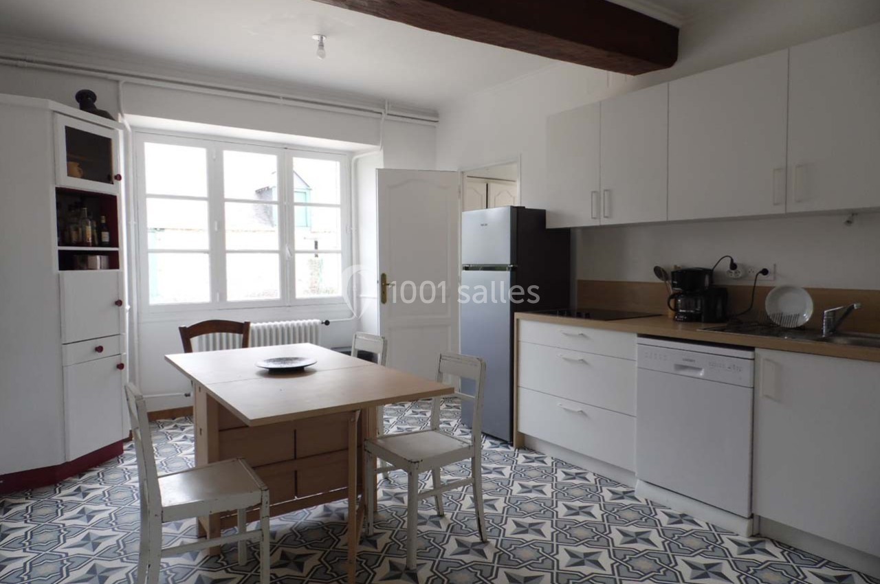 Cuisine lumineuse avec meubles blancs, table en bois, réfrigérateur, lave-vaisselle et sol en carreaux à motifs.