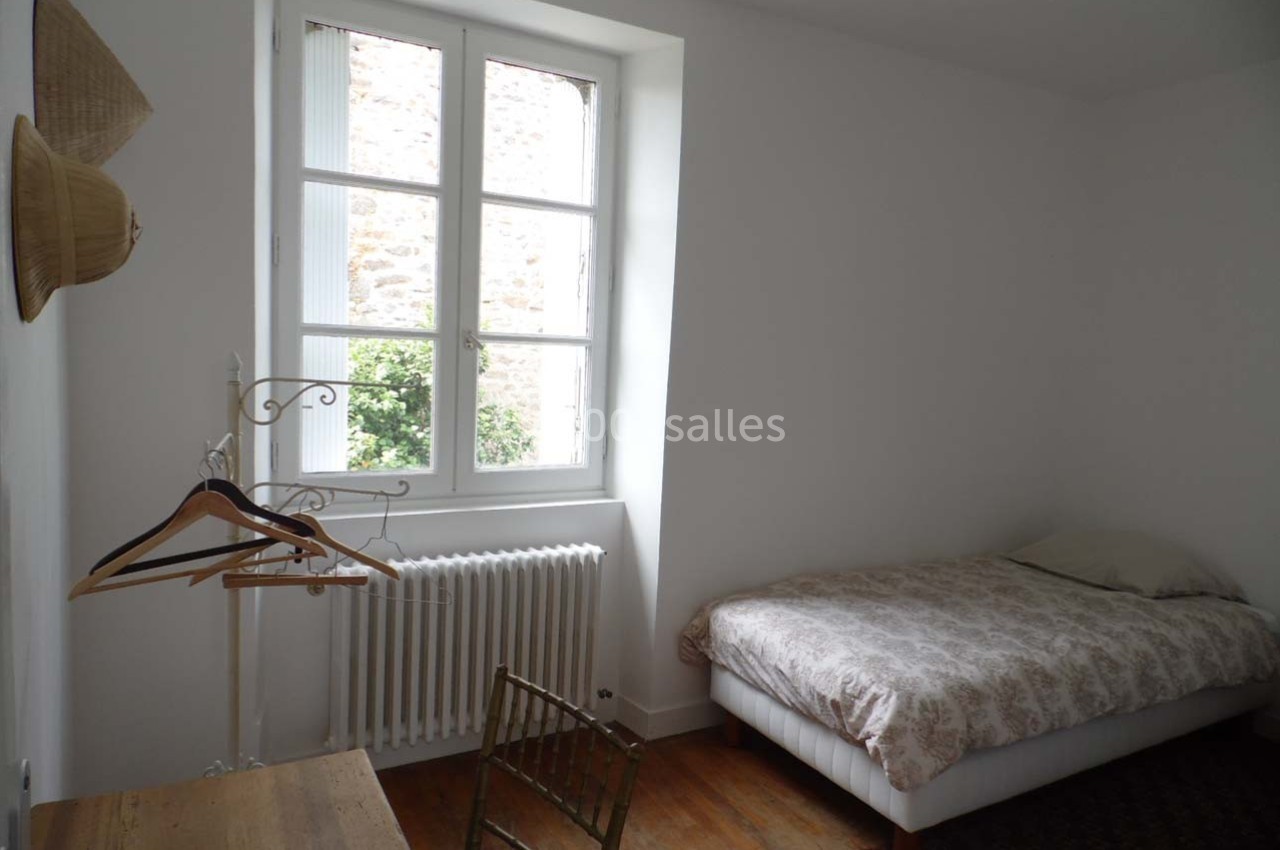 Chambre lumineuse avec lit simple, bureau en bois, chaise, porte-manteaux et fenêtre donnant sur un mur en pierre.