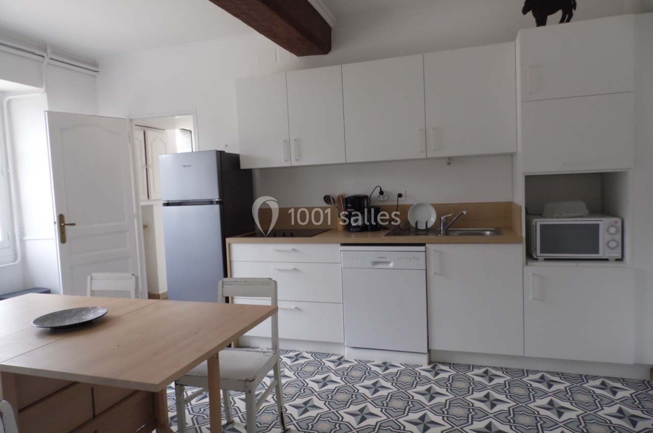 Cuisine lumineuse avec meubles blancs, réfrigérateur, micro-ondes, lave-vaisselle et table en bois sur sol à motifs.