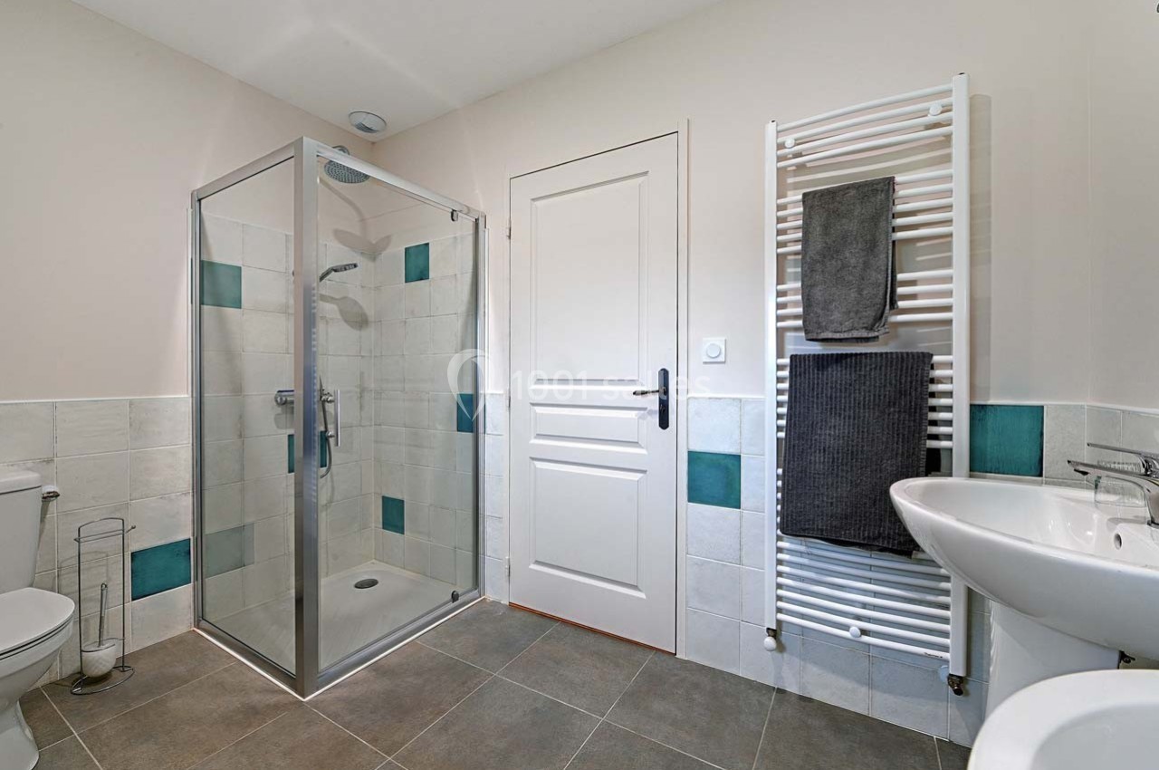 Salle de bain avec douche vitrée, lavabo, toilettes, sèche-serviettes et carrelage aux tons neutres et turquoise.