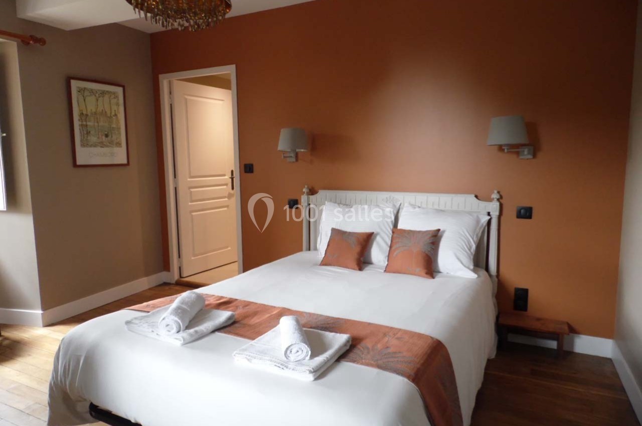 Chambre avec lit double, linge blanc, coussins et jeté de lit orange, murs peints en orange et éclairage mural.