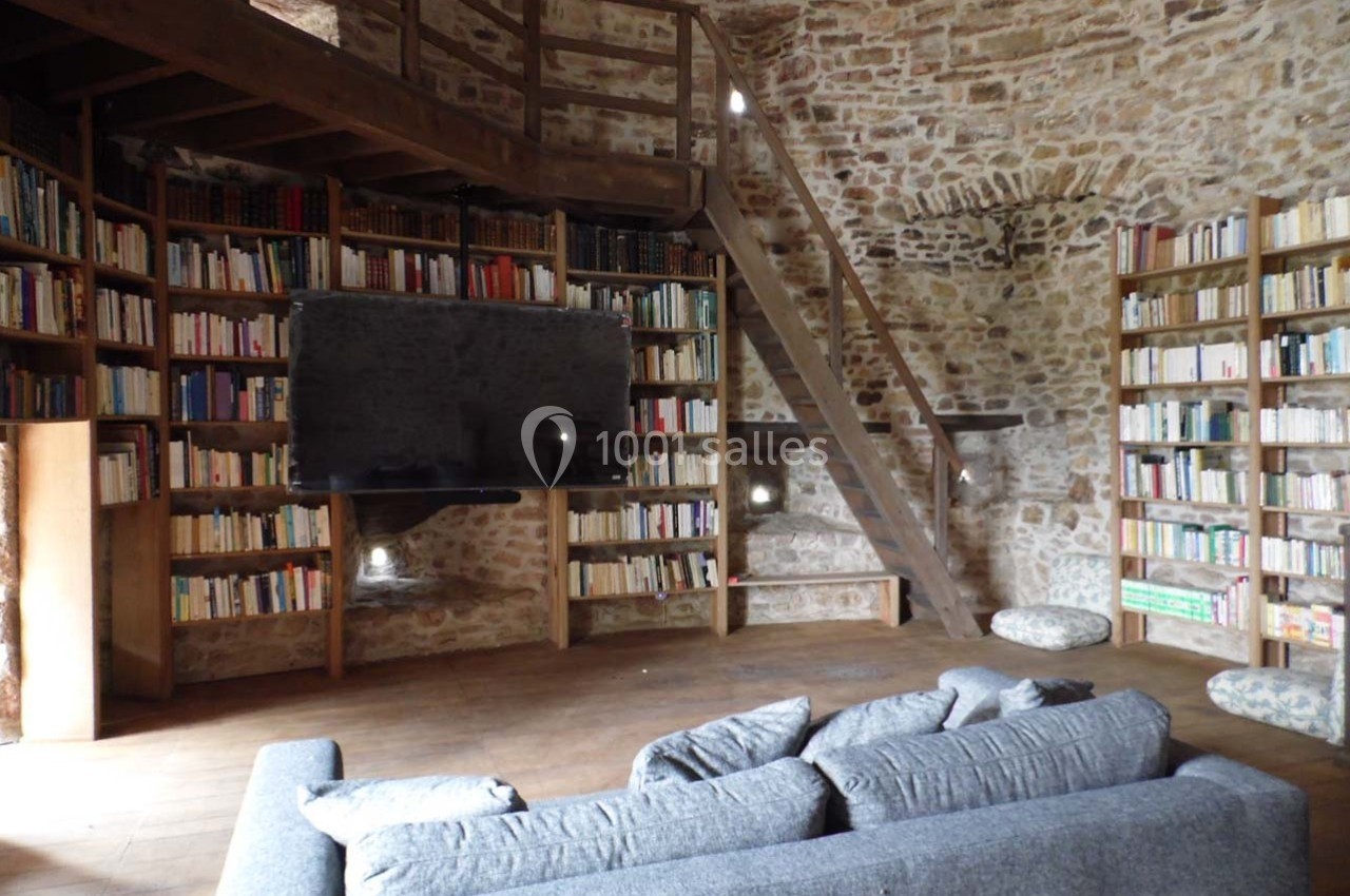 Bibliothèque avec murs en pierre, étagères remplies de livres, canapé gris et escalier en bois menant à une mezzanine.