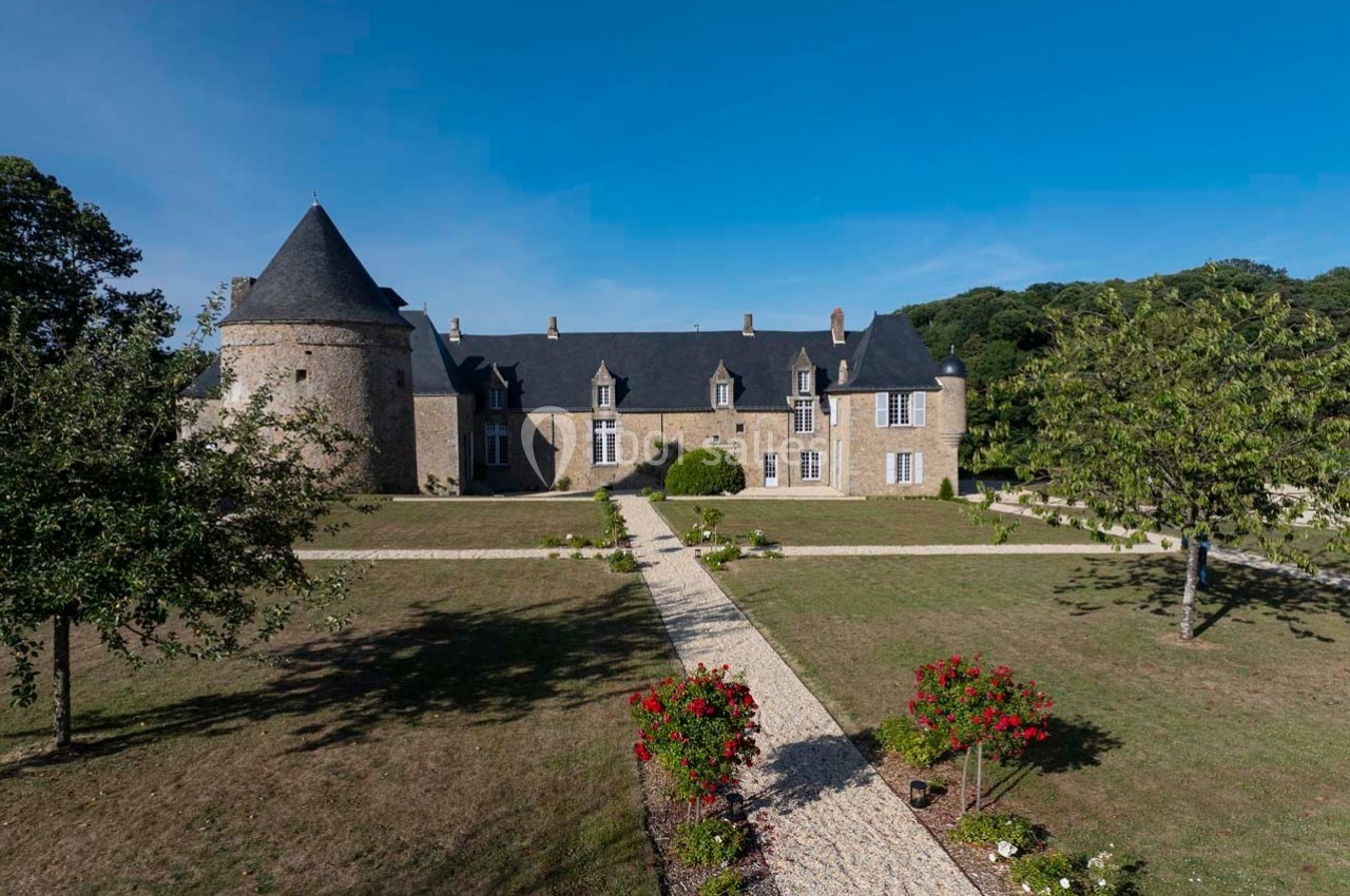 Château en pierre avec une tour ronde, entouré d'un jardin arboré et d'allées pavées sous un ciel bleu.