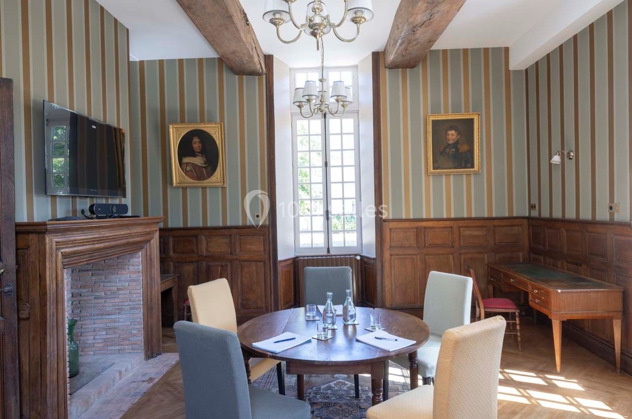 Salle de réunion lumineuse avec table ronde, chaises colorées, cheminée, poutres apparentes et portraits accrochés au mur.