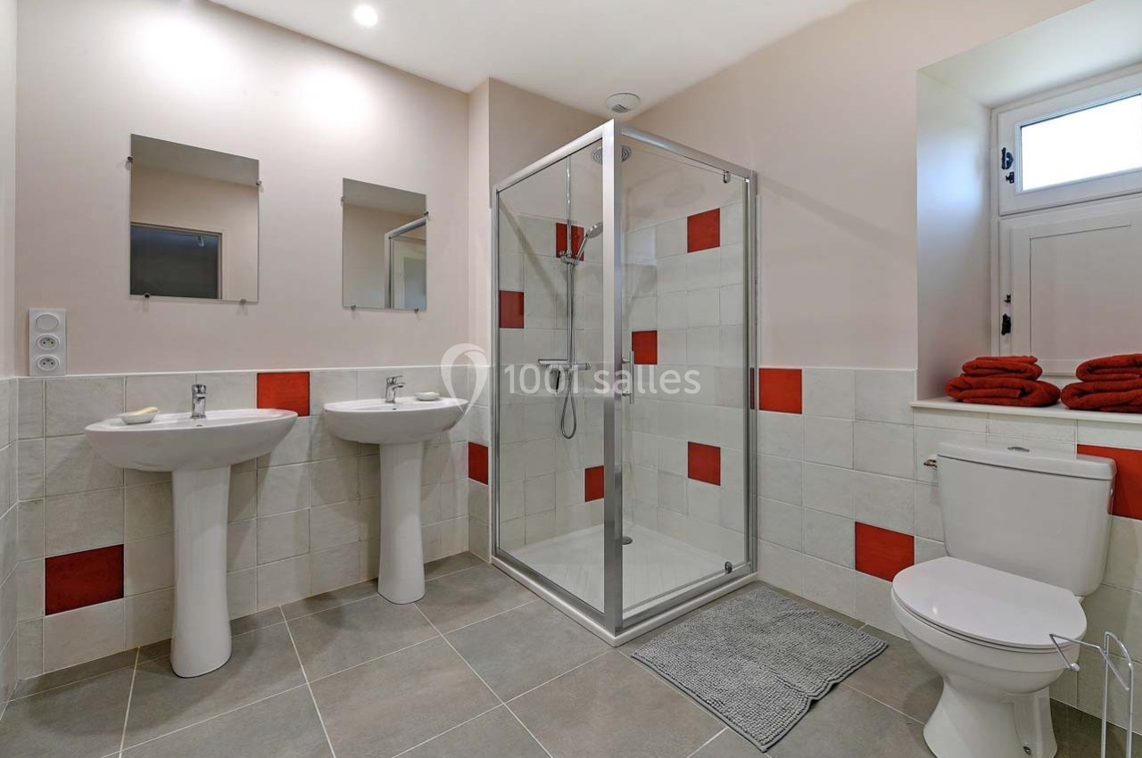 Salle de bain moderne avec douche vitrée, deux lavabos, toilettes et serviettes rouges pliées sur une étagère.