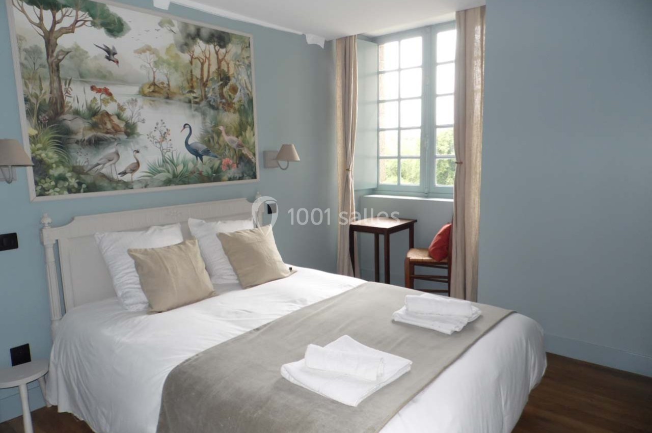 Chambre lumineuse avec un lit double, des coussins beiges, une grande fenêtre et une décoration murale représentant des…