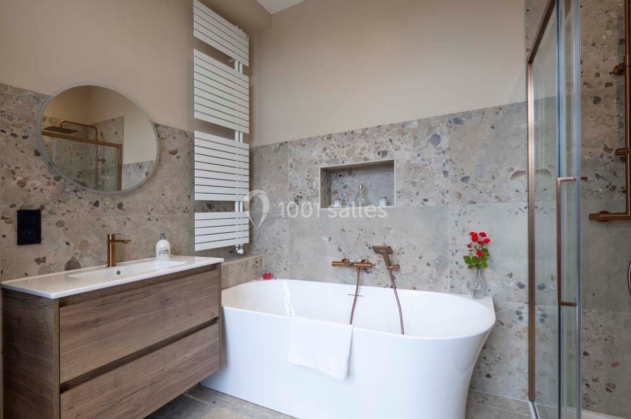 Salle de bain moderne avec baignoire blanche, meuble en bois, miroir rond et carrelage effet pierre.