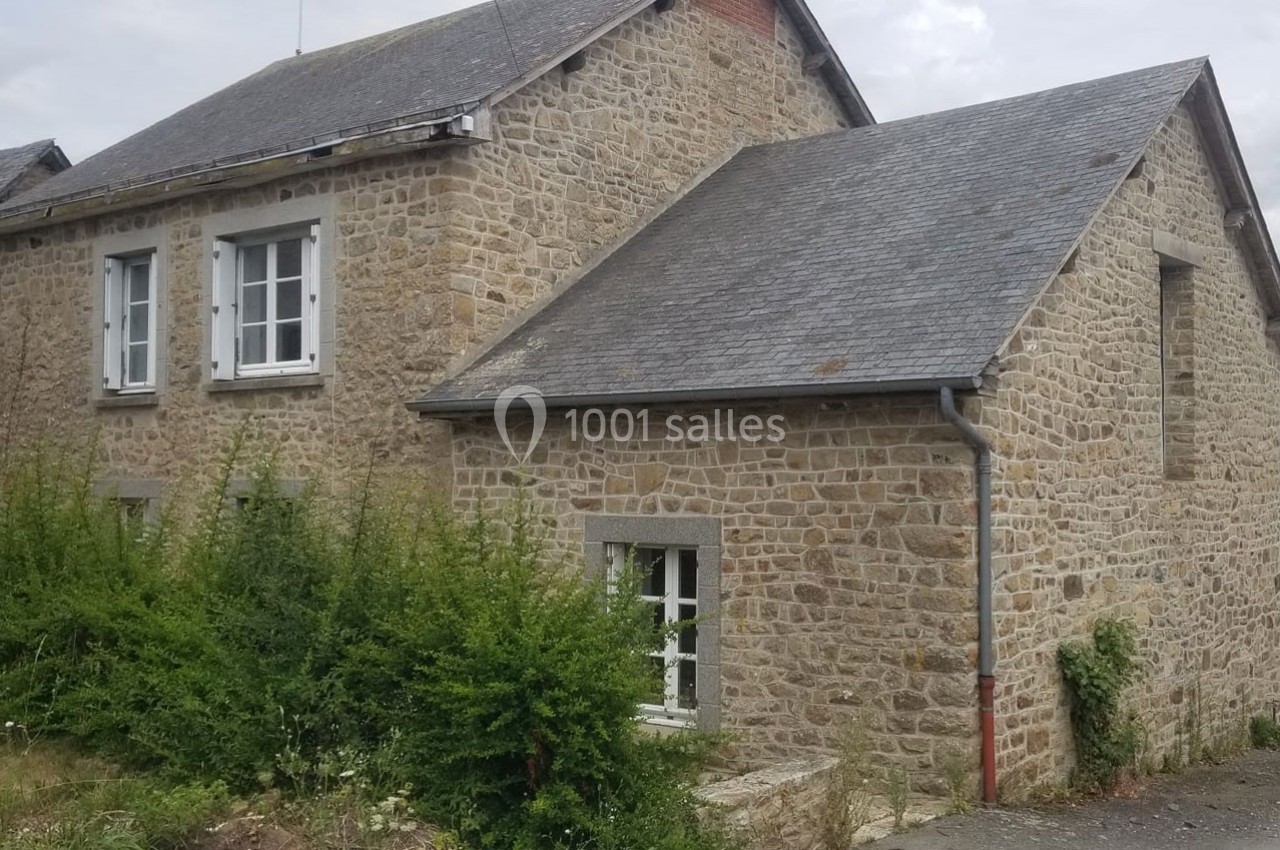 Maison en pierre avec toit en ardoise, fenêtres blanches et végétation dense à l'avant.