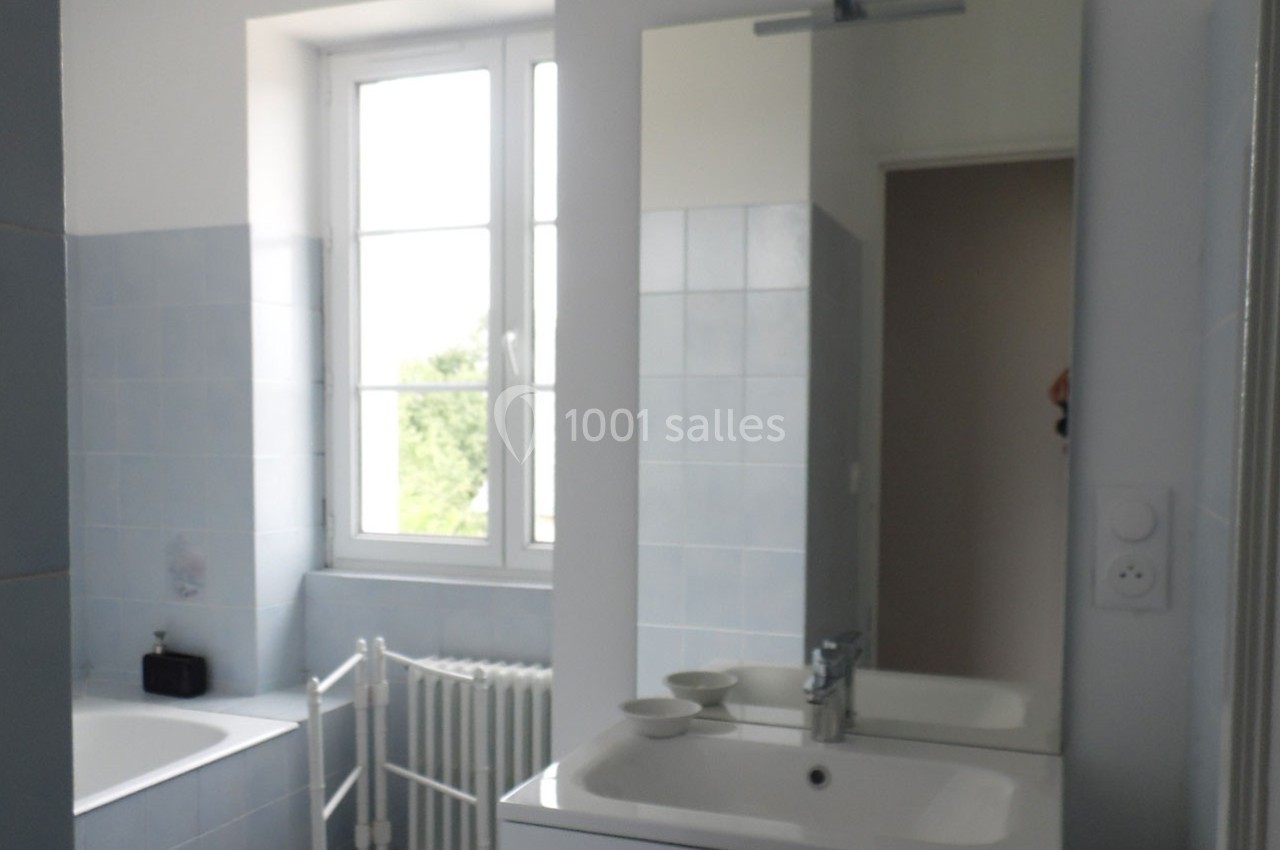 Salle de bain lumineuse avec carrelage bleu clair, lavabo, miroir, baignoire et fenêtre donnant sur l'extérieur.