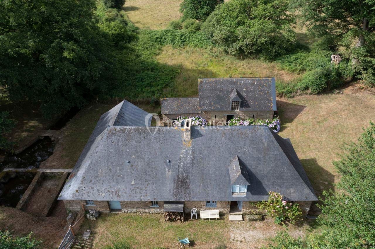 Vue aérienne d'une maison en pierre avec toit en ardoise, entourée de verdure et de champs.
