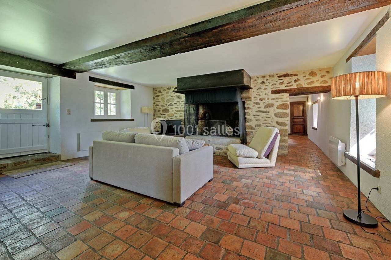 Salon rustique avec murs en pierre, cheminée imposante, canapé beige, fauteuils et sol en carreaux de terre cuite.