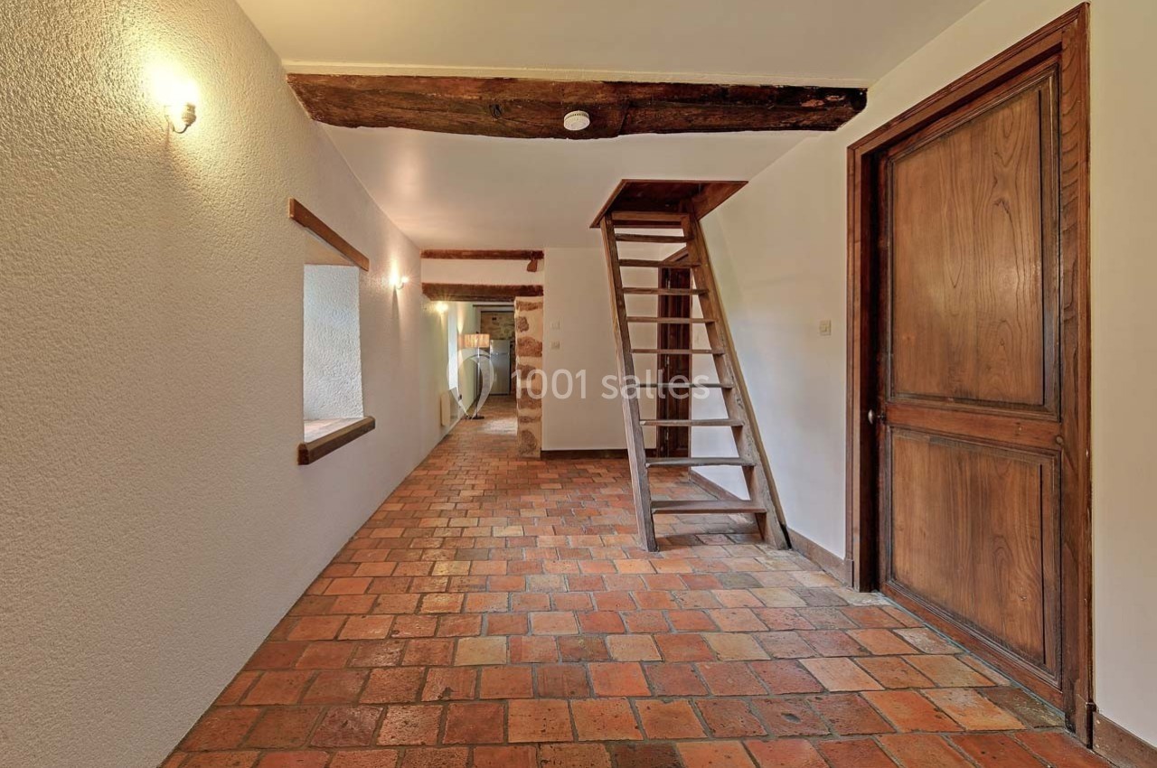 Escalier en bois dans un couloir avec sol en carreaux de terre cuite, murs clairs et poutres apparentes.