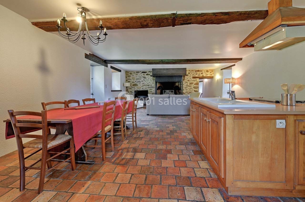 Salle à manger avec table en bois, cuisine ouverte et salon avec cheminée, sur un sol en carreaux de terre cuite.