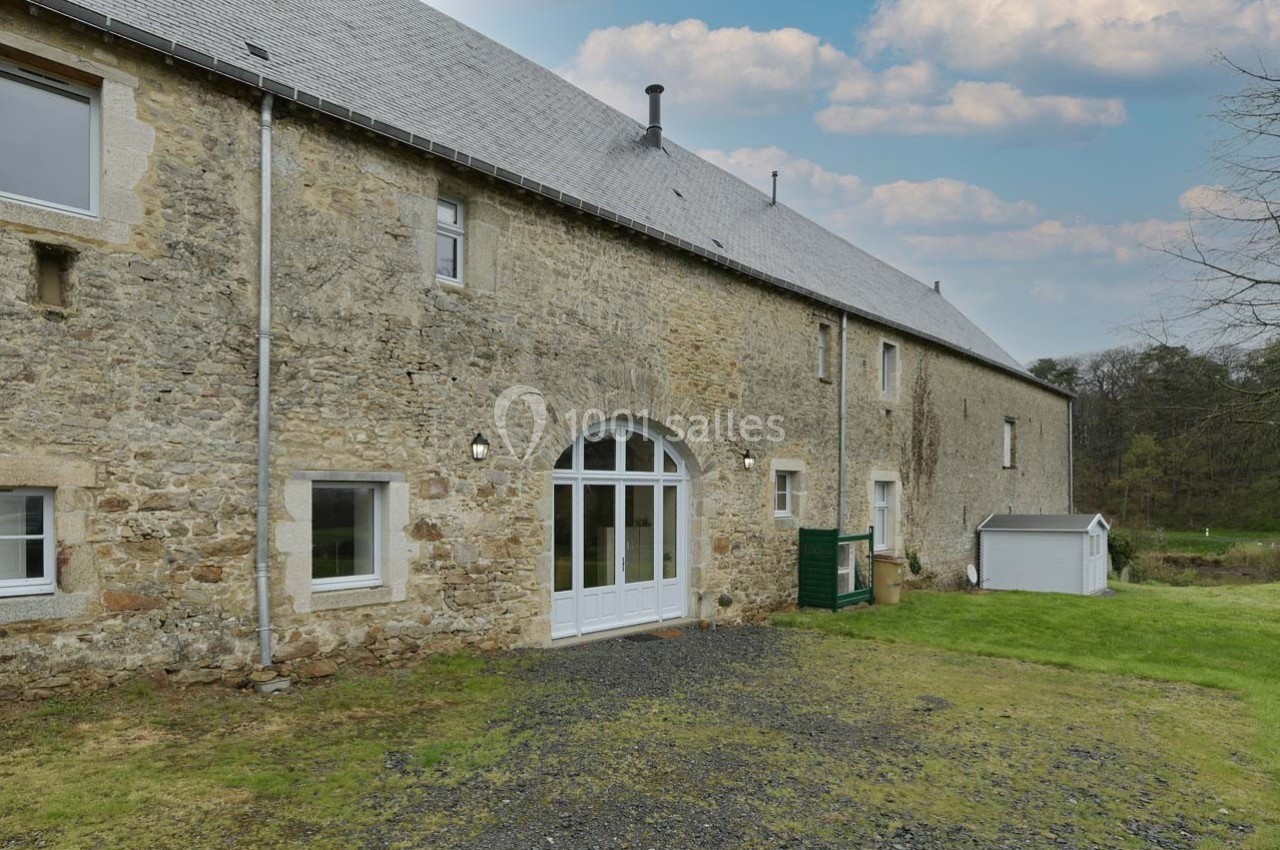 Façade en pierre d'une grande bâtisse avec porte vitrée, entourée de pelouse et d'arbres sous un ciel partiellement nuageux.