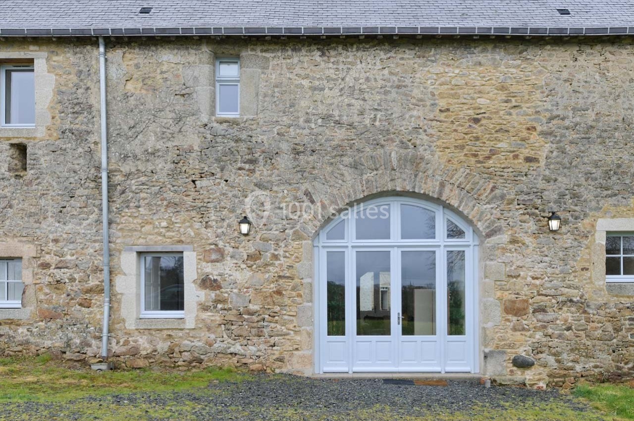 Façade en pierre d'une maison avec une grande porte vitrée cintrée et des fenêtres, entourée de pelouse et de gravier.