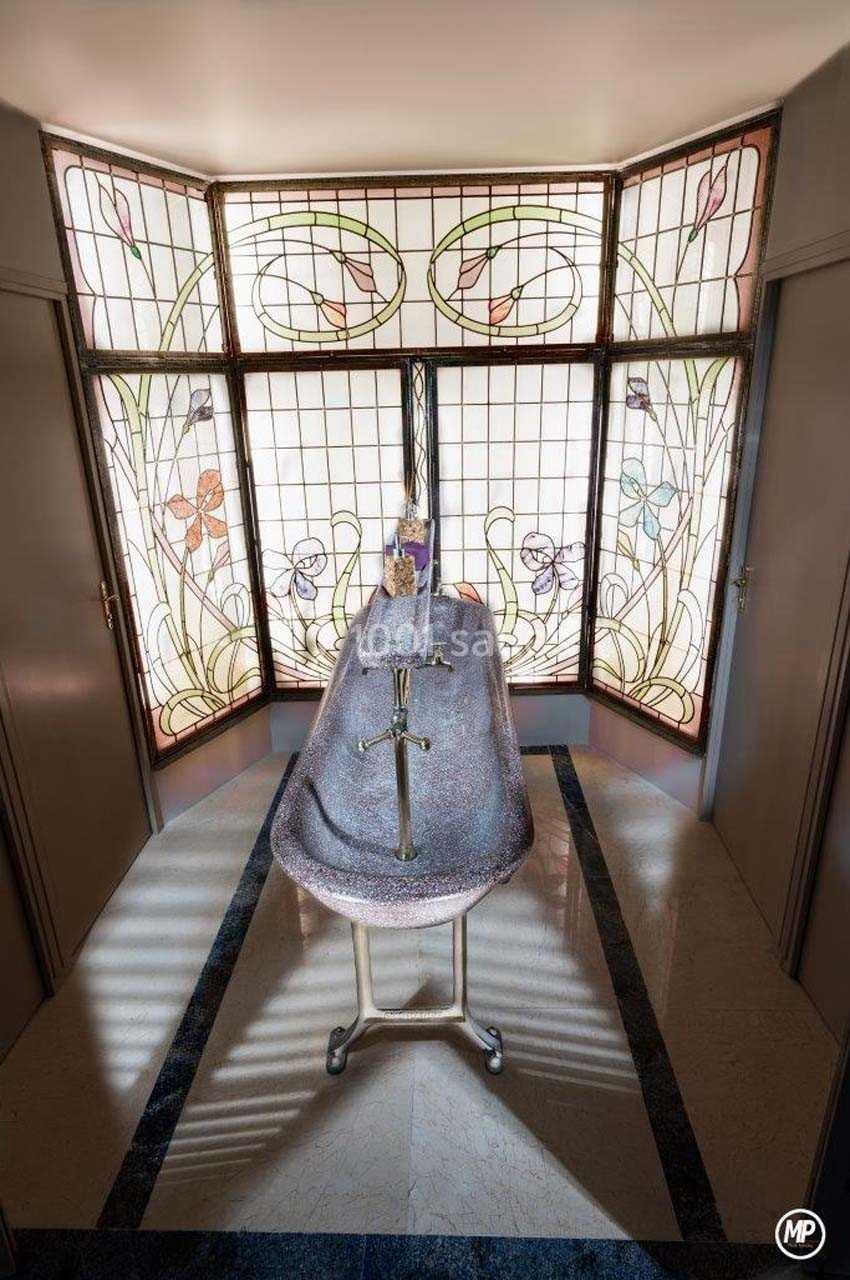 Un cercueil recouvert d'un drap gris avec une croix, placé devant un vitrail décoratif dans une pièce lumineuse.