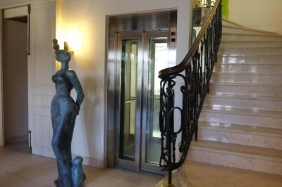 Statue moderne près d'un escalier en marbre et d'un ascenseur à portes vitrées dans un hall lumineux.