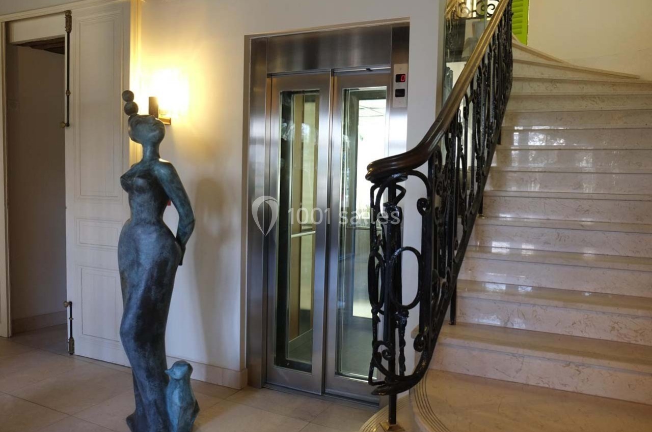 Statue moderne près d'un escalier en marbre et d'un ascenseur à portes vitrées dans un hall lumineux.