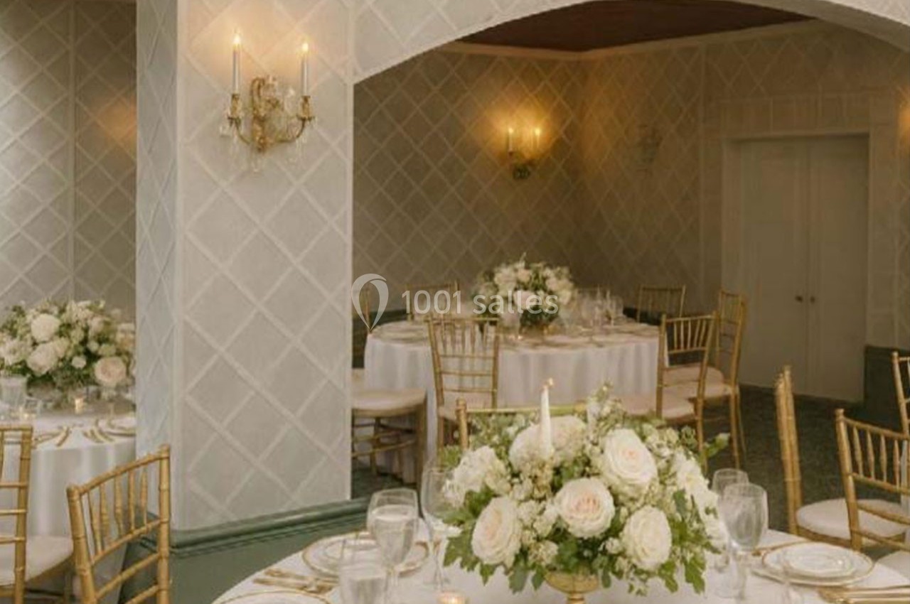 Salle élégante décorée pour un événement, avec tables rondes, chaises dorées et compositions florales blanches.