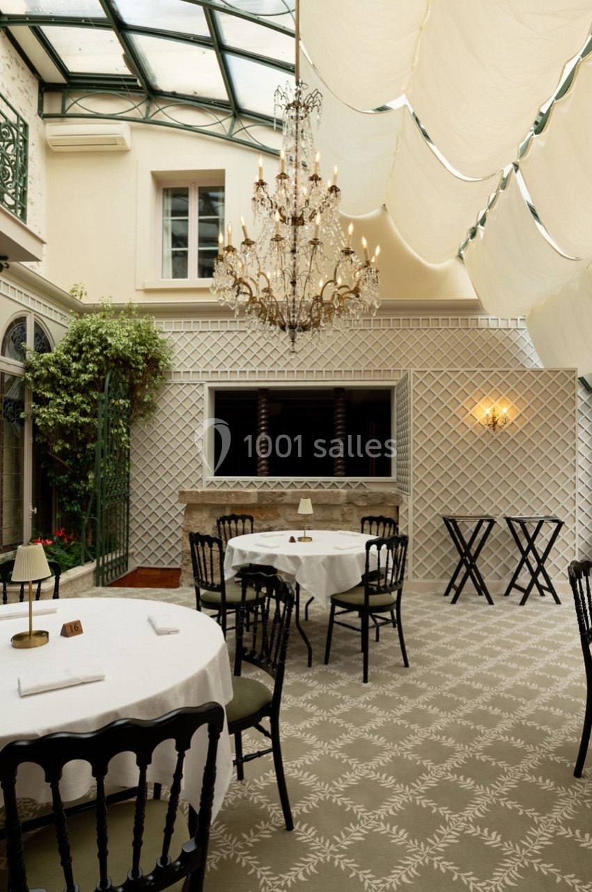 Salle lumineuse avec verrière, tables dressées, chaises noires et lustre central dans un décor élégant et classique.