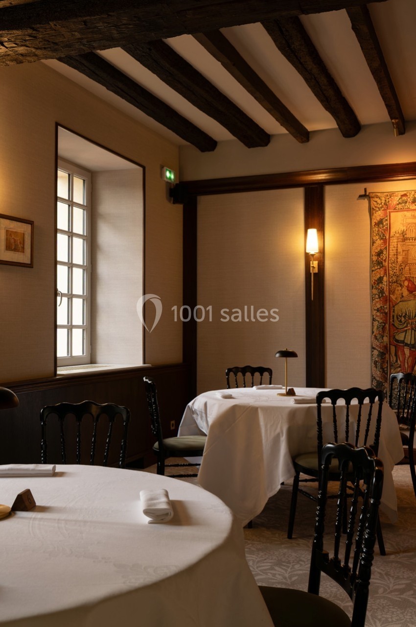 Salle de restaurant élégante avec tables dressées, chaises noires, poutres apparentes et lumière tamisée.
