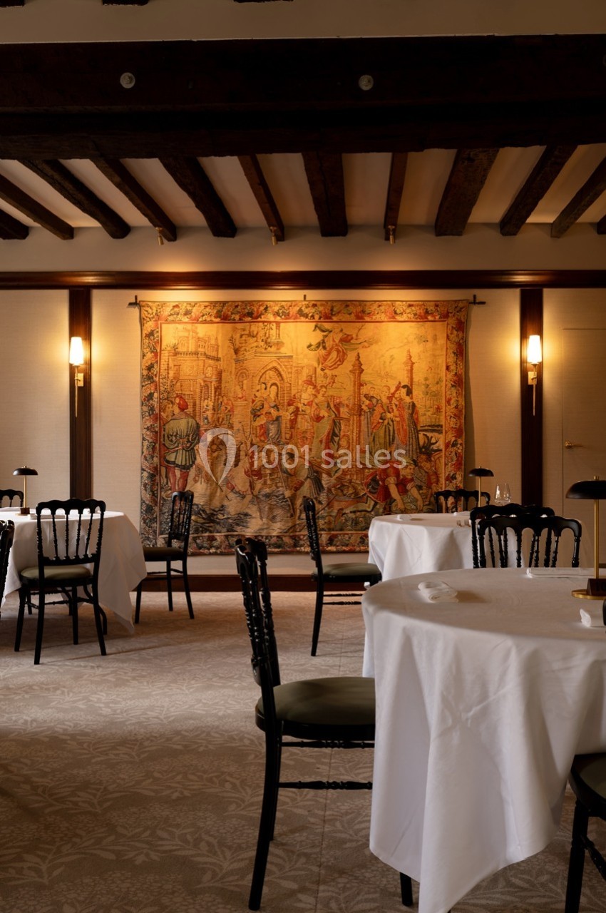 Salle de restaurant élégante avec tables dressées, chaises noires et une grande tapisserie murale colorée en arrière-plan.