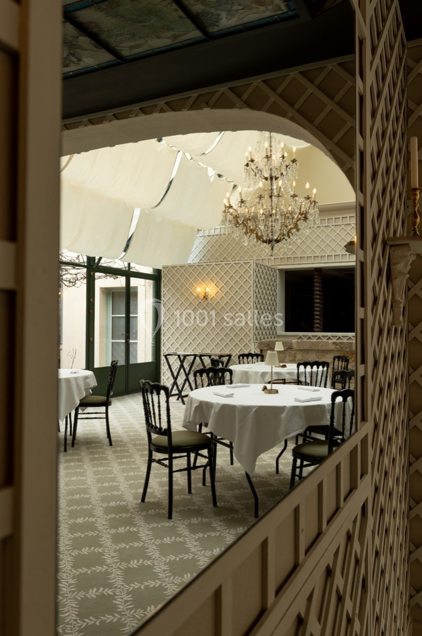 Salle de restaurant élégante avec tables dressées, chaises noires, lustre en cristal et plafond partiellement vitré.